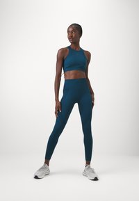 Sostén deportivo en color verde azulado y leggings de tiro alto hechos de tejido elástico, con paneles texturizados. La modelo lleva zapatillas deportivas grises.