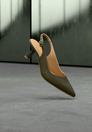 Πράσινο παπούτσι slingback με kitten τακούνι, μυτερή μύτη και λουρί με πόρπη, κρεμασμένο πάνω από μια γκρι επιφάνεια με θολό φόντο.
