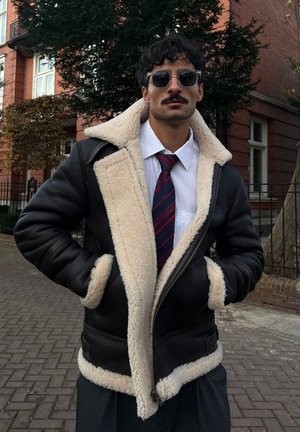 Brown leather jacket with a cream shearling lining, featuring a zipper closure, large collar, and ribbed cuffs. Striped tie underneath.