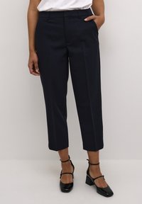 Pantalons taillés bleu marine avec une coupe droite, longueur mi-mollet, plis à l'avant et passants de ceinture, associés à des talons en cuir verni noir.