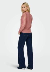 Felpa a maniche lunghe in rosa polveroso con maniche trasparenti e ricamate con fiori. Abbinata a jeans a gamba larga blu scuro.