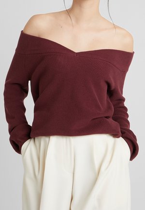 Femme portant un pull en maille bordeaux à épaules dénudées, les mains dans les poches d'un pantalon blanc ample taille haute, sur fond gris.