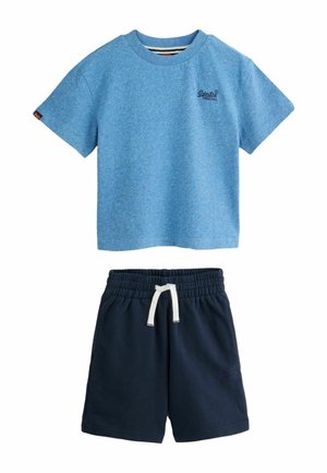Barn med skulderlangt hår står iført en lyseblå t-shirt, mørke shorts med hvid snor, mærkesokker og beige sneakers.