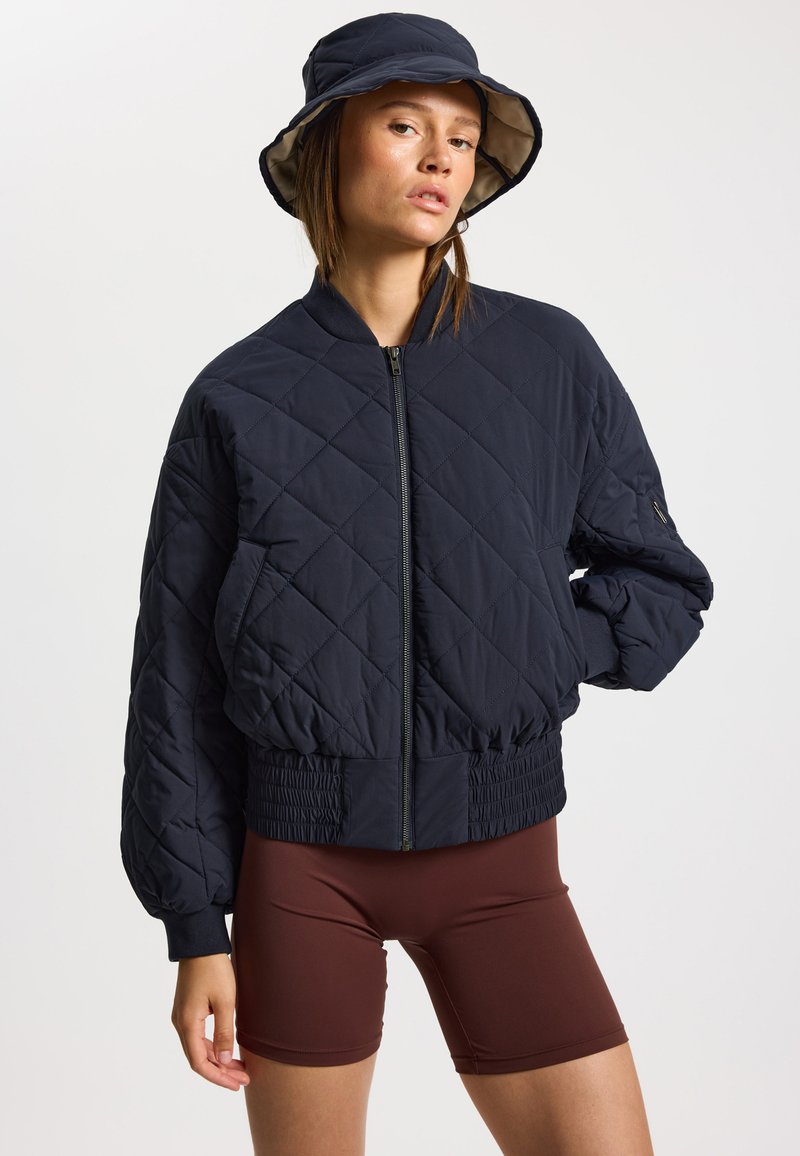 Veste bomber matelassée bleu marine avec ceinture et poignets élastiques, dotée d'une fermeture éclair. Comprend également un chapeau bob coordonné bleu marine.