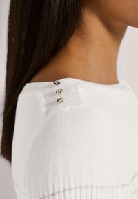 Weißes Strickoberteil mit einem runden Ausschnitt und drei dekorativen goldenen Knöpfen an der Schulter. Der Stoff hat eine weiche Textur.