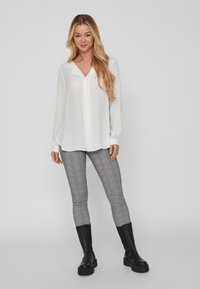 Weiße Bluse mit langen Ärmeln, V-Ausschnitt und Knopfleiste, kombiniert mit grauen karierter Hose mit strukturiertem Muster und schwarzen klobigen Stiefeln.