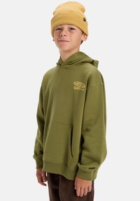 Olívazöld kapucnis pulóver elülső zsebből és arany színű Quiksilver logóból. Mustár sárga kötött sapkával viselve. Sima szövettextúra.