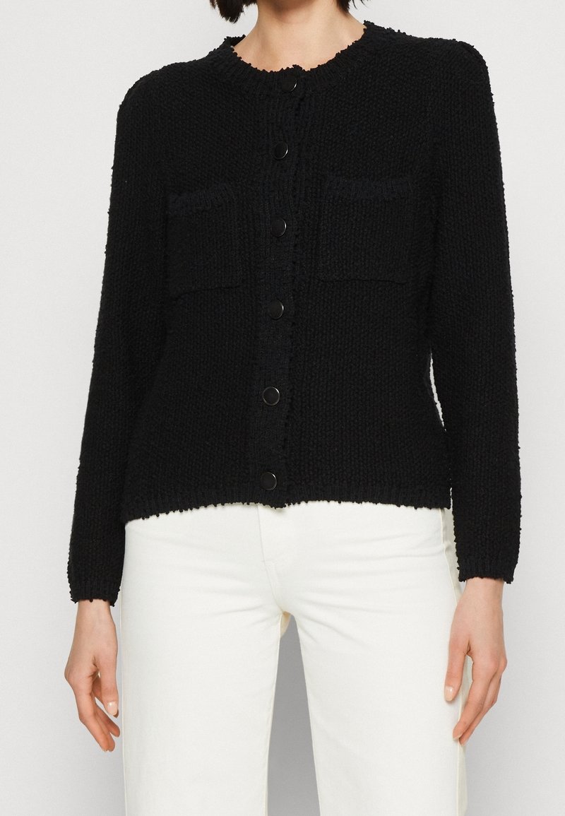 Cardigan noir texturé à boutons, manches longues et deux poches avant, porté avec un pantalon blanc, montré des épaules aux hanches.