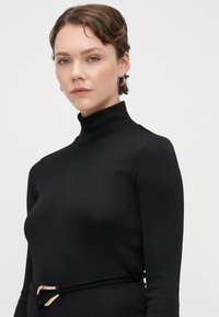 Schwarzes Rollkragenoberteil mit einer taillierten Passform, geripptem Material und einem schmalen Gürtel mit einer goldenen Schnalle als Akzent. Das Model hat eine kurze Frisur.