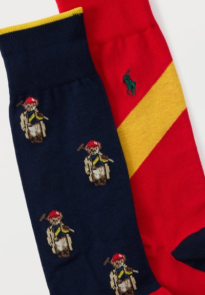 Δύο πολύχρωμες κάλτσες, η μία σε ναυτικό μπλε με μοτίβα καβαλάρηδων πόλο που φορούν κόκκινες καπέλες, και η άλλη κόκκινη με μια κίτρινη διαγώνια λωρίδα και πράσινο λογότυπο.