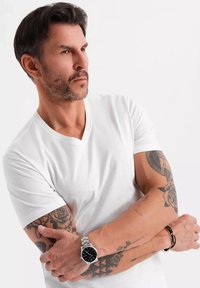 T-shirt bianco a scollo a V, maniche corte. Mostra di tatuaggi sulle braccia, orologio argento con quadrante nero, capelli scuri pettinati all'indietro. Sfondo neutro.