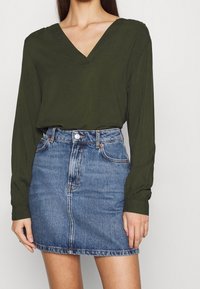 Blouse olive verte à manches longues avec un col en V, associée à une jupe en denim bleu clair dotée d'une fermeture à boutons et de poches avant.