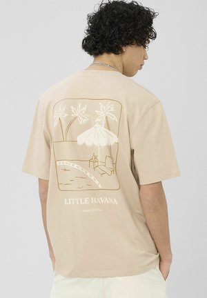 Homme aux cheveux bouclés portant un t-shirt beige montrant un imprimé au dos avec des palmiers, un parasol de plage, une chaise longue et le texte « Little Havana Miami, Florida ».