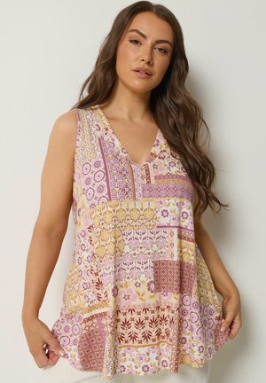 Kobieta o długich brązowych włosach, ubrana w bezrękawnik z dekoltem w serek w patchworkowy wzór w odcieniach różu, żółci i czerwieni, trzymająca się za krawędź bluzki.