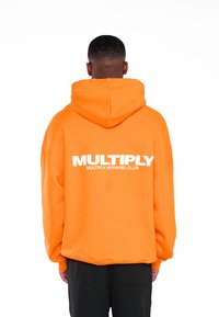 Sudadera oversized naranja con un gran logo blanco en la parte trasera. Cuenta con capucha ajustable y puños de canalé. Textura de tela suave.