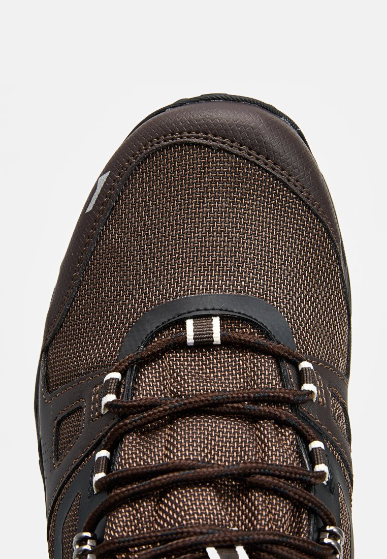 Zapato de textil marrón con acentos sintéticos, que presenta un diseño robusto, superficie texturizada y un sistema de cordones distintivo.