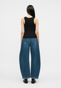 Γυναίκα που στέκεται με την πλάτη στην κάμερα, φοράει μαύρο τοπ χωρίς μανίκια, φαρδιά μπλε τζιν και μαύρα παπούτσια πλατφόρμα σε απλό φόντο.