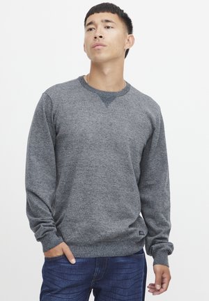 Grå genser med manchetter og lange ermer, ribbestruktur ved halsen og mansjettene, samt et subtilt mørkt mønster. Kombinert med blå jeans.