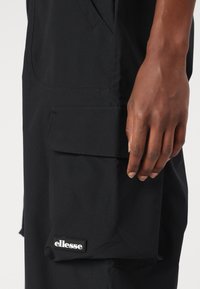 Crne cargo hlače od glatkog materijala, s bočnom džepom s preklopom i brendiranim logom "ellesse" blizu rubnog dijela.