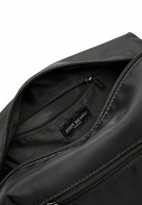 Zwarte nylon tas interieur met een gladde textuur, voorzien van een ritszak en een label met de tekst "John Henric Sweden." Lineaire stiksels zijn zichtbaar.