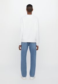 White sweatshirt with ribbed cuffs, paired with light blue straight-leg jeans. Casual footwear is white. The background is plain.