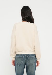 Ανοιχτό μπεζ φούτερ με στρογγυλή λαιμόκοψη και χαλαρούς ώμους, συνδυασμένο με ανοιχτόχρωμα τζιν, που παρουσιάζει άνετη εφαρμογή και ριμπ μανσέτες.