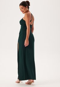 Abito lungo verde realizzato in tessuto morbido, con corpino aderente, dettagli arricciati e uno spacco alto fino alla coscia. Sandali con tacco trasparente completano il look.