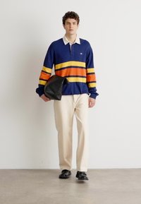 Marine rugby polo skjorte med oransje og gule striper, kombinert med beige bukser. Holder en svart, teksturert clutchveske. Svart sko.