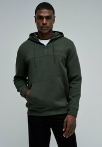 Sudadera con cremallera de color verde oscuro, con un bolsillo tipo canguro, tejido texturizado y logo bordado en el pecho. Combinada con pantalones negros.
