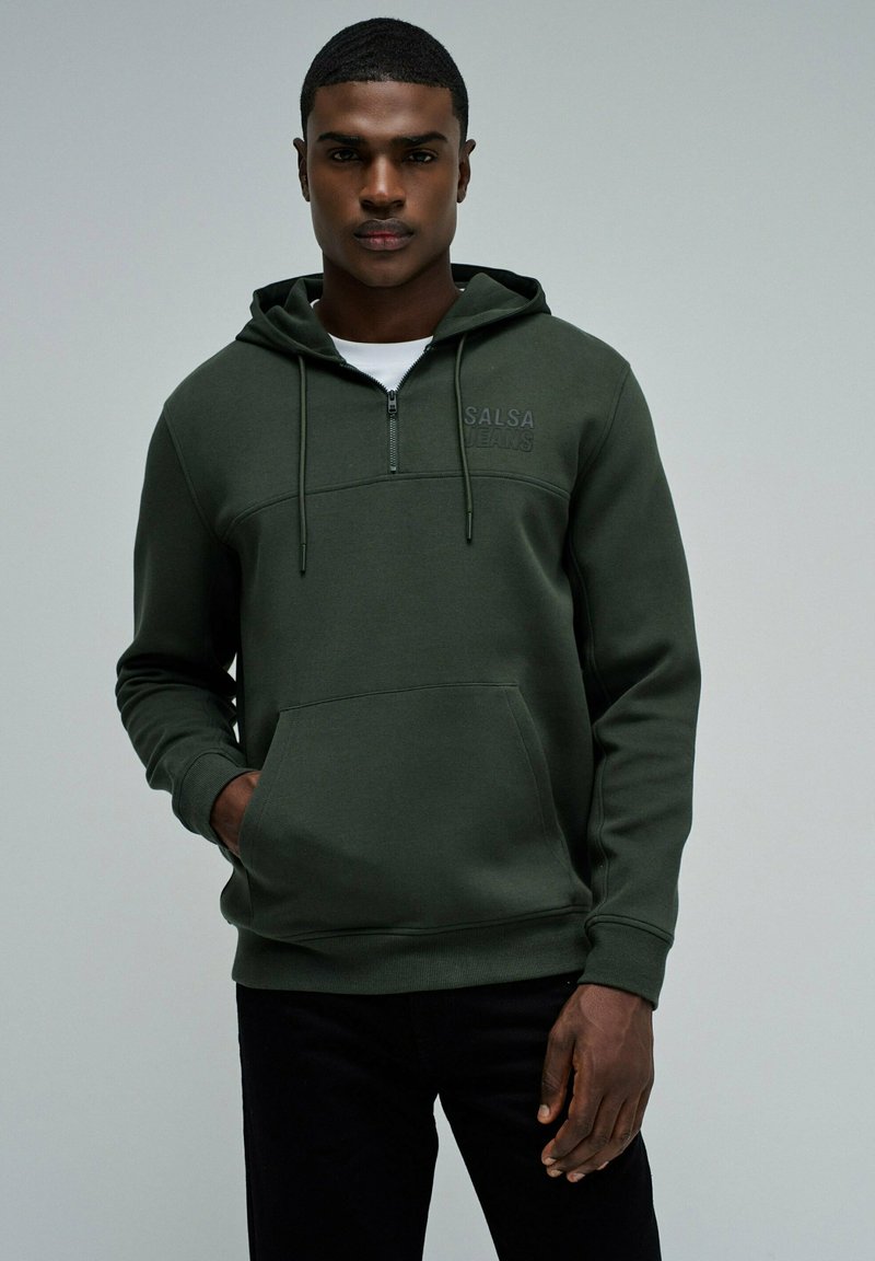Sudadera con cremallera de color verde oscuro, con un bolsillo tipo canguro, tejido texturizado y logo bordado en el pecho. Combinada con pantalones negros.