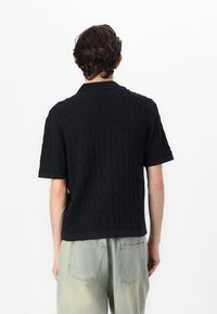 Polo shirt noir en tricot avec un motif en vagues, manches courtes et col. Associé à un pantalon en denim vert clair.