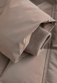 Bézs színű, bélelt dzseki steppelt mintával és a hajtott pulóvermandzsetta belülről látható. Simább, könnyű anyag minimális részletezéssel.