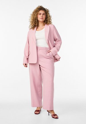 Žena s kučeravými vlasmi stojí oblečená v svetloružovom obleku, bielom topu a bordových otvorených lodičkách na bielej pozadí.