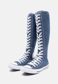 Blaue Canvas-Stiefel bis zur Kniehöhe mit weißen Ösen und Schnürsenkeln, schwarzem gestreiften Gummi-Sohle und kreisförmigem Logo-Patch an der Seite.
