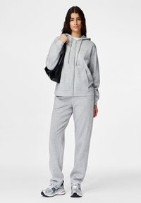 Woman standing wearing light gray zip-up hoodie and matching sweatpants, holding black shoulder bag, and wearing silver sneakers.