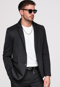 Homme portant un blazer noir, un t-shirt blanc, un pantalon foncé, des lunettes de soleil et un collier chaîne argenté, debout avec une main dans la poche devant un fond uni.