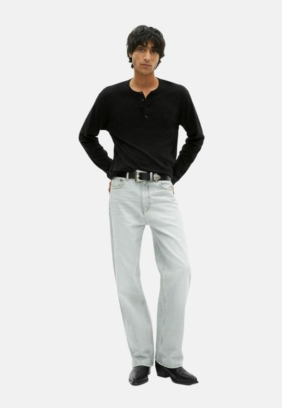 Jeune homme aux cheveux bruns portant un tee-shirt longue manche henley noir, un jean bleu clair, une ceinture noire et des chaussures noires, se tenant debout avec les mains sur les hanches.