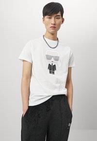Balta medvilninė T-shirt su animaciniu personažu, apsirengusiu kostiumu ir saulės akiniams. Suderinta su juodomis švelniomis kelnėmis su šoninėmis kišenėmis ir logotipo detale.