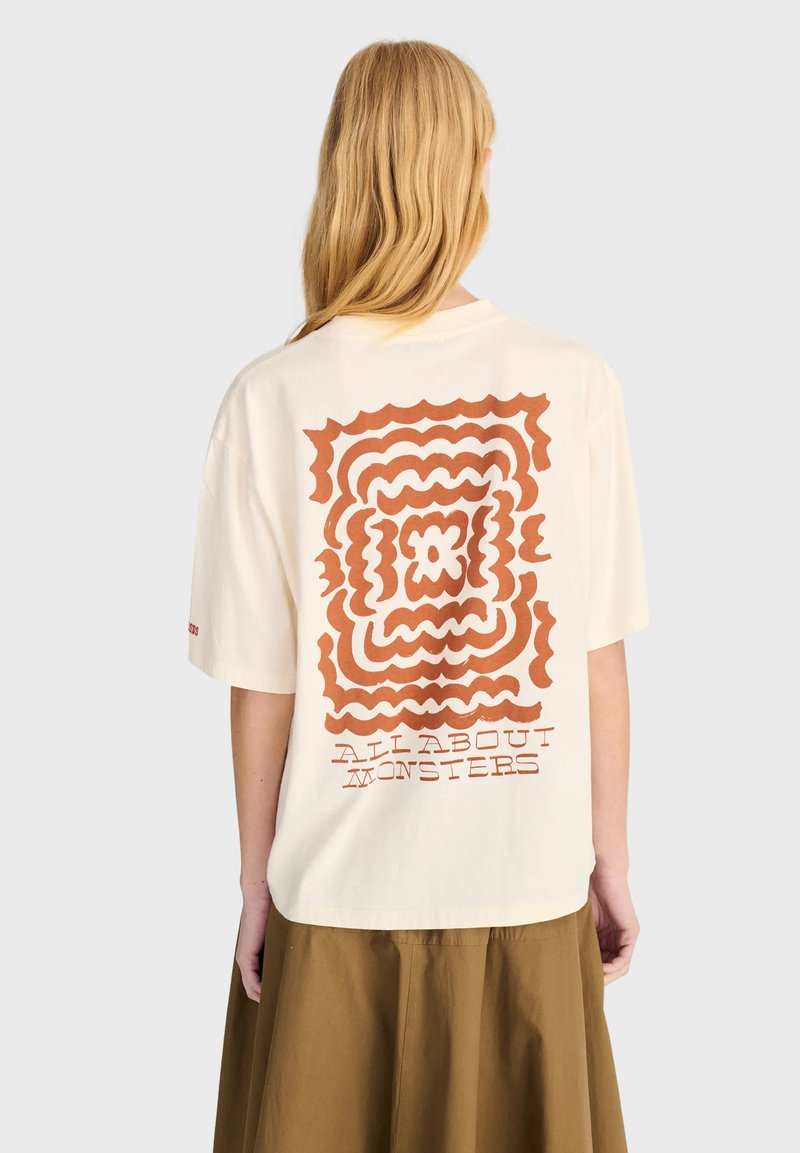 Personne aux longs cheveux blonds, portant un t-shirt beige clair avec un motif carré ondulé marron et le texte "All About Monsters" dans le dos.