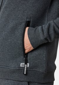 Grå zip-up hoodie i mjukt material, med en sidoficka med svart dragkedja och en etikett med texten "DENIM FACTORY" i nederkant.