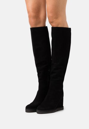 Botas de ante negro por encima de la rodilla con tacón de cuña, con cremallera lateral y acabado sin costuras. Textura suave y diseño elegante.