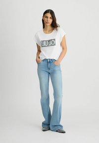 Bílý krátký rukáv T-shirt s nápisem "LIU.JO" v černých flitrech, spárovaný se světle modrými zvonovými džíny a černobílými vzorovanými botami.
