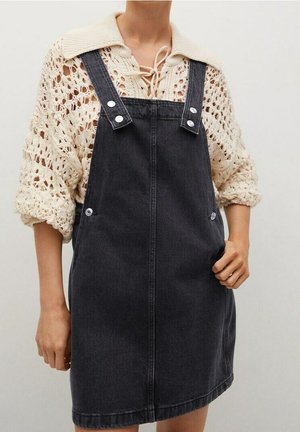 Robe salopette en denim noir à coupe droite, avec bretelles réglables, boutons argentés en accent, et un pull en crochet doux de couleur crème en dessous.