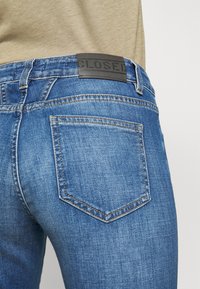 Close-up of back pocket on blue denim jeans worn with beige top, showing black leather brand patch on waistband.