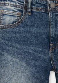 Jean bleu denim avec texture délavée, couture orange, poche avant, passants de ceinture, bouton en métal et fermeture éclair visibles.