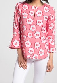 Blouse fleurie rose avec des accents blancs et rouges, dotée d'une encolure fendue et de manches cloche, confectionnée dans un tissu léger. Associée à un pantalon blanc.