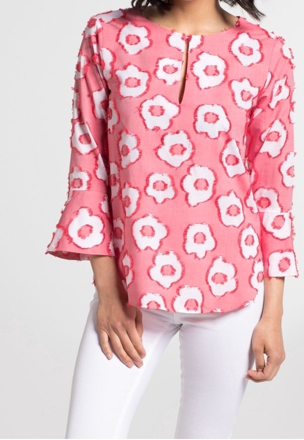 Blouse fleurie rose avec des accents blancs et rouges, dotée d'une encolure fendue et de manches cloche, confectionnée dans un tissu léger. Associée à un pantalon blanc.
