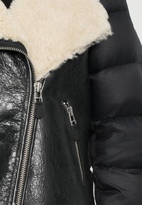 Schwarze Lederjacke mit einem cremefarbenen Kunstschafskragen. Sie hat eine glatte, glänzende Textur und silberne Reißverschlüsse auf der Vorderseite.