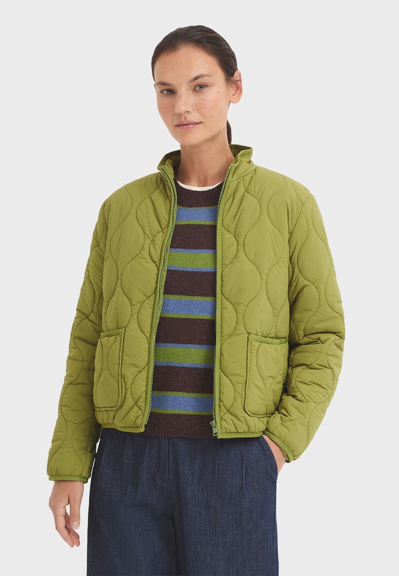 Gequilted groene jacket met een opstaand kraag, twee voorkzakken en een rits. Draag het over een gestreept sweater en donkere broek.