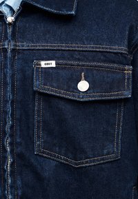 Τζιν μπουφάν σε σκούρο μπλε με φερμουάρ στο κλείσιμο, δύο μπροστινές τσέπες, ασημένιες μεταλλικές λεπτομέρειες και αντίθετη κίτρινη ραφή. Ετικέτα "OBEY" στην τσέπη.